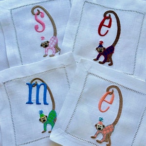 May include: Four white linen napkins with embroidered monkeys holding letters. Each monkey is wearing a party hat and a different colored outfit. The letters are S, E, M, and E.