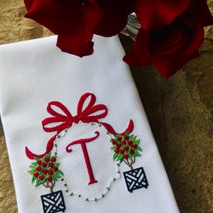 May include: White decorative towel with a red embroidered bow, the letter "T", and two small potted plants with red berries. A bouquet of red roses is in the background.