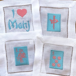 May include: Four white linen napkins with embroidered designs. The first napkin has a pink heart and the text "I love Mahj". The second napkin has a blue square with a pink Chinese character. The third napkin has a blue square with a pink bird and the number "1". The fourth napkin has a blue square with the number "5" and five pink circles.