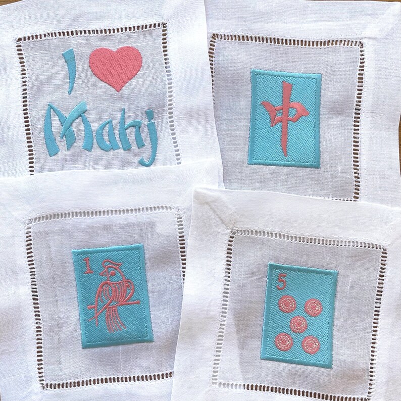 May include: Four white linen napkins with embroidered designs. The first napkin has a pink heart and the text "I love Mahj". The second napkin has a blue square with a pink Chinese character. The third napkin has a blue square with a pink bird and the number "1". The fourth napkin has a blue square with the number "5" and five pink circles.