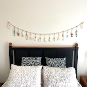 Op de afbeelding: Een slaapkamer met een bed met witte gewatteerde beddengoed, kussens met patronen en een zwart hoofdbord. Boven het bed hangt een decoratieve slinger van touw met strandthema-ornamenten, waaronder zeesterren, surfplanken en het woord "hello".