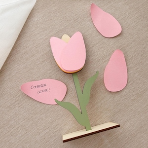 Puede incluir: Un soporte de flores de madera en forma de tulipán con pétalos de flores de papel rosa y hojas. Una nota adhesiva rosa con el texto "COMPRAR LECHE!" está pegada al soporte.