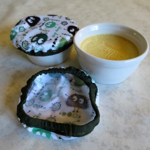 Op de afbeelding: Twee witte keramische kommen met groene en zwarte gepatenteerde stoffen deksels. Een kom bevat een gele, custard-achtige substantie.