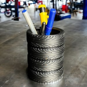 May include: A black tire-shaped pencil holder with four stacked tires. The holder is filled with pens and pencils, including a yellow pen, a blue pen, and a white pen.