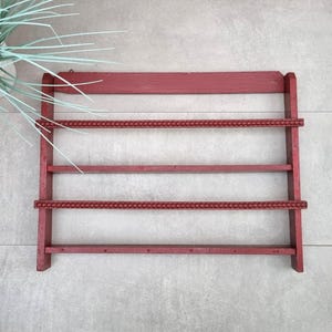Può includere: Un portapiatti in legno rosso con quattro ripiani orizzontali. I ripiani superiori e inferiori sono lisci, mentre i ripiani centrali hanno un bordo decorativo con perline. Il portapiatti è montato su una parete grigio chiaro.
