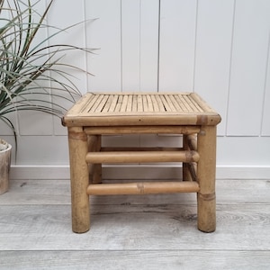 Op de afbeelding: Een kleine, vierkante bamboe kruk met een lattenbodem en drie horizontale steunbalken. De natuurlijk gekleurde houten kruk heeft vier stevige poten en is geschikt voor binnen of buiten. De kruk is ongeveer 30 cm hoog.