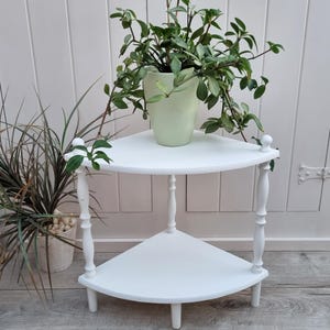 Op de afbeelding: Witte hoekplank met twee niveaus, met gedraaide houten poten en een plant in een lichtgroene pot op de bovenste plank.