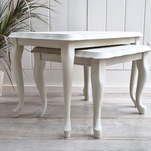 May include: A set of two white nesting tables with a distressed finish. The tables have a simple, elegant design with four legs each.
