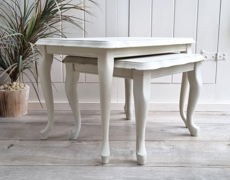 May include: A set of two white nesting tables with a distressed finish. The tables have a simple, elegant design with four legs each.
