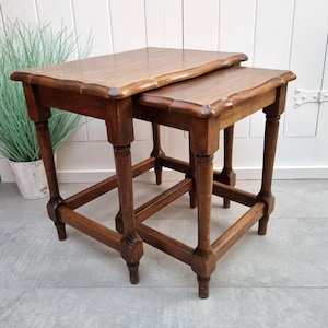 Può includere: Un set di due tavolini a incastro antichi in legno. I tavolini sono di colore marrone caldo con un bordo smerlato. Le gambe sono tornite e collegate da supporti inferiori. I tavolini sono di diverse dimensioni, progettati per incastrarsi.