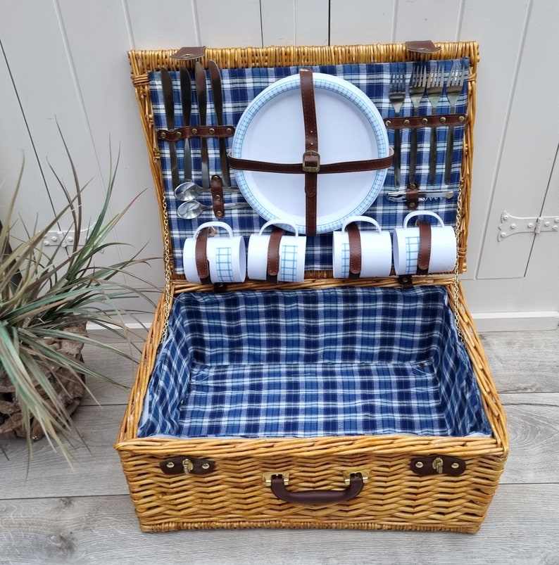 May include: A wicker picnic basket with a blue and white checkered lining. The basket is open and contains a white plate, four mugs, and silverware.