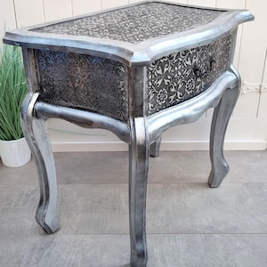 Op de afbeelding: Zilverkleurige bijzettafel met een bloempatroon op het blad en de lade. De tafel heeft gebogen poten en een enkele lade met een donkere knop. De lade en het blad hebben een zwart-zilveren bloemenmotief. Een decoratief meubelstuk.