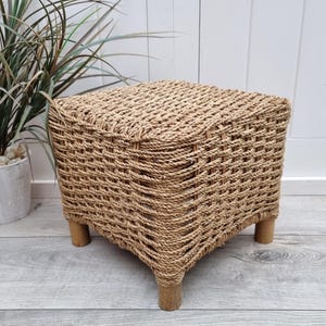 Puede incluir: Un taburete cuadrado tejido de hierba marina con patas de madera natural. El taburete está hecho de un material tejido natural y tiene un color marrón claro.