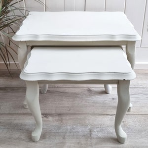 May include: A set of two white nesting tables with scalloped edges and turned legs. The tables are made of wood and have a distressed finish.