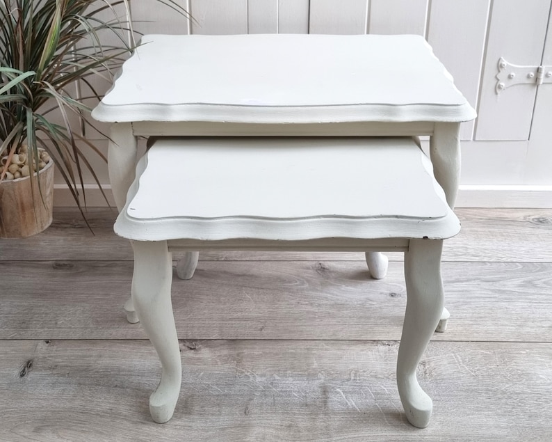 May include: A set of two white nesting tables with scalloped edges and turned legs. The tables are made of wood and have a distressed finish.