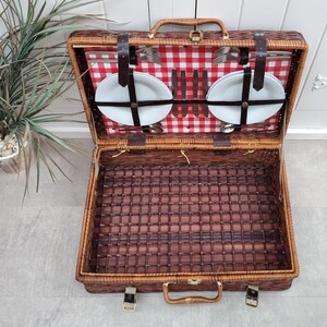 May include: A brown wicker picnic basket with a red and white checkered lining. The basket is open and contains two white plates, silverware, and a set of knives.