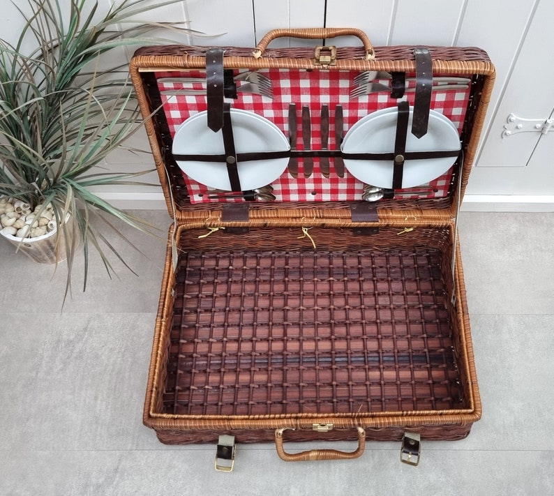 May include: A brown wicker picnic basket with a red and white checkered lining. The basket is open and contains two white plates, silverware, and a set of knives.
