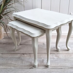 May include: A set of two white nesting tables with a scalloped edge and turned legs. The tables are made of wood and have a distressed finish.