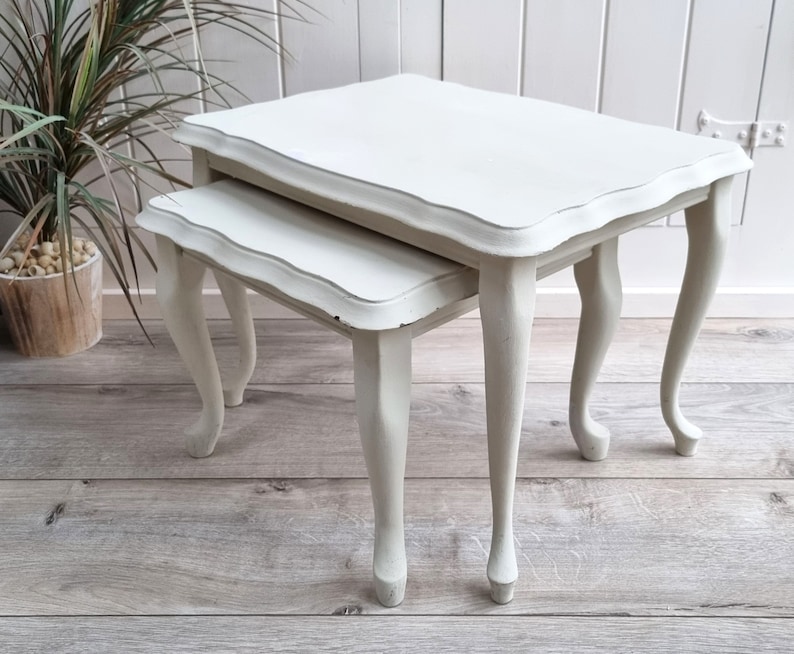 May include: A set of two white nesting tables with a scalloped edge and turned legs. The tables are made of wood and have a distressed finish.