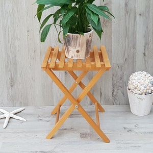 Op de afbeelding: Een houten opvouwbare plantenstandaard met een natuurlijke afwerking. Een plant in een pot met groene bladeren staat op de standaard. De standaard staat op een lichte houten vloer.