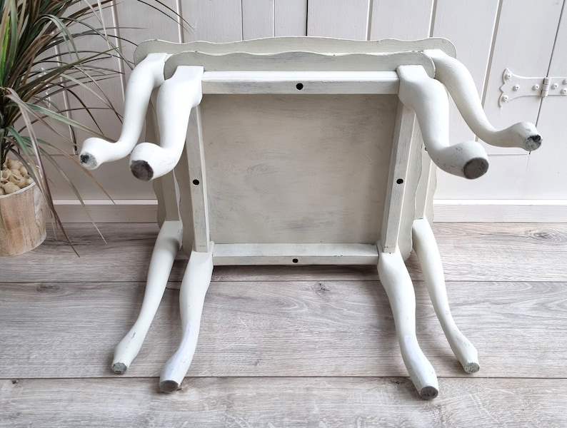 May include: White painted wooden side table with a scalloped edge and four legs. The table is distressed and has a rustic look.