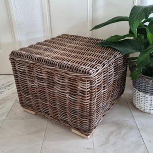 Könnte beinhalten: Ein quadratischer, flacher Hocker aus geflochtenem braunem Weidengeflecht. Das Möbelstück verfügt über eine flache Oberseite und wird von vier kleinen, hellen Holzfüßen an der Basis gestützt, was eine natürliche, strukturierte Optik erzeugt.