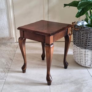 Op de afbeelding: Een kleine, vierkante houten bijzettafel met een donkerbruine afwerking. De tafel heeft vier gebogen poten en een eenvoudig, elegant ontwerp. De tafel is ongeveer 51 cm hoog en 46 cm breed.