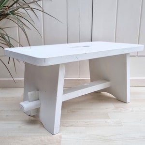May include: A white wooden stool with a simple design. The stool has a rectangular top and four legs. The legs are connected by a horizontal support beam.