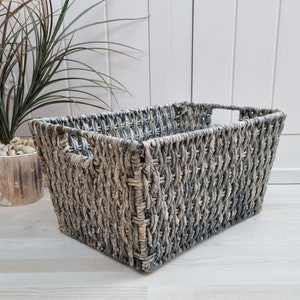Puede incluir: Una cesta de almacenamiento tejida gris con forma rectangular y un patrón tejido. La cesta está hecha de materiales naturales y tiene un aspecto rústico.