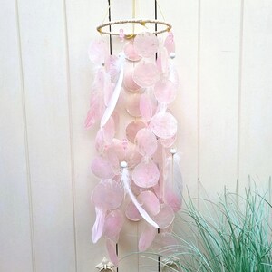 Op de afbeelding: Een roze en witte windgong met een goudkleurige ring aan de bovenkant. De windgong heeft roze, cirkelvormige schelpachtige stukken en witte veren die aan de ring hangen. De windgong hangt voor een witte houten muur.
