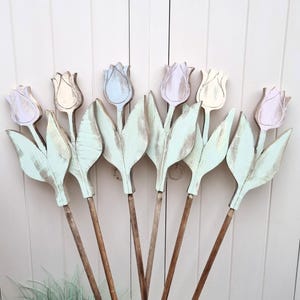 May include: Wooden tulip decorations with pastel-colored petals and mint green leaves. The tulips are mounted on long, brown wooden stems. The background is a white wooden door, creating a rustic, spring-themed display.