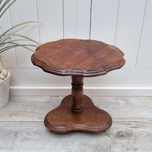 May include: A small, round, wooden table with a scalloped edge top and a single, turned pedestal base. The table is made of dark brown wood and has a rustic, antique look.