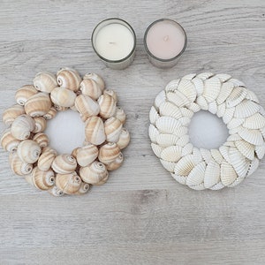 May include: Two seashell wreaths, one made of brown and white shells and the other made of white shells, with two candles in the background.