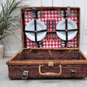 May include: A brown wicker picnic basket with a red and white checkered lining. The basket is open and contains two white plates, silverware, and a set of knives.