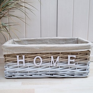 May include: A whitewashed wicker storage basket with a linen liner. The basket has the word "HOME" spelled out in white letters on the front.