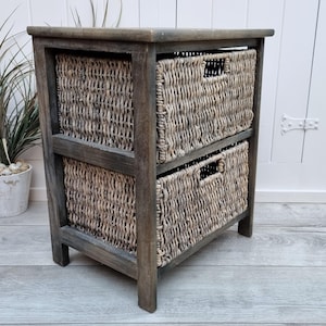Puede incluir: Una unidad de almacenamiento de madera de dos niveles con cestas tejidas. La unidad está pintada de color gris oscuro y tiene un acabado desgastado. Las cestas están hechas de materiales naturales y tienen un color marrón claro.