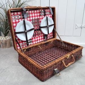 May include: A wicker picnic basket with a red and white checkered lining. The basket is open and contains two white plates, silverware, and a variety of other picnic essentials.