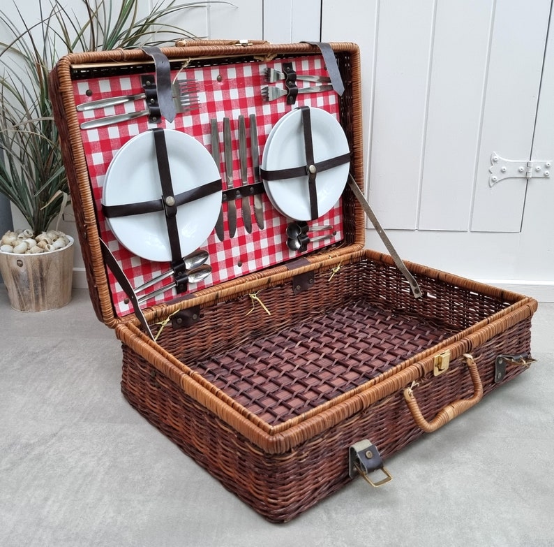 May include: A wicker picnic basket with a red and white checkered lining. The basket is open and contains two white plates, silverware, and a variety of other picnic essentials.