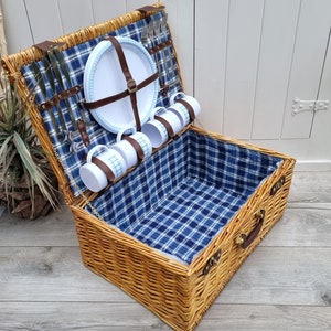 May include: A wicker picnic basket with a blue and white plaid lining. The basket is open and contains white ceramic mugs, plates, and silverware.