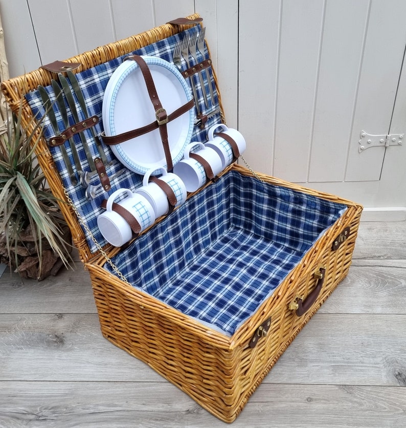 May include: A wicker picnic basket with a blue and white plaid lining. The basket is open and contains white ceramic mugs, plates, and silverware.