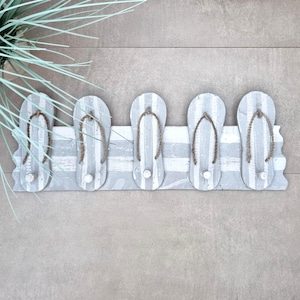 Peut inclure: Un porte-manteau mural décoratif en forme de tongs. Il comporte cinq crochets en forme de tongs, peints en gris et blanc avec des rayures. Les lanières sont en corde. Idéal pour une décoration sur le thème de la plage.
