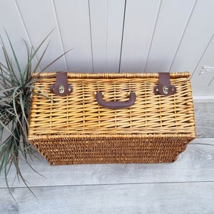 May include: A woven wicker picnic basket with a brown leather handle and brass clasps. The basket is closed and has a natural brown color.