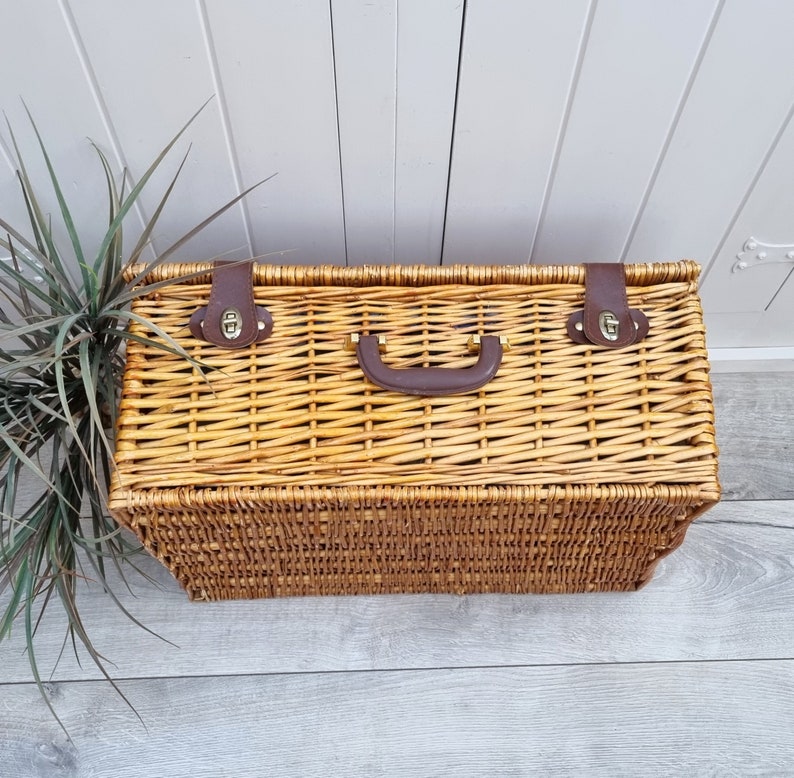 May include: A woven wicker picnic basket with a brown leather handle and brass clasps. The basket is closed and has a natural brown color.