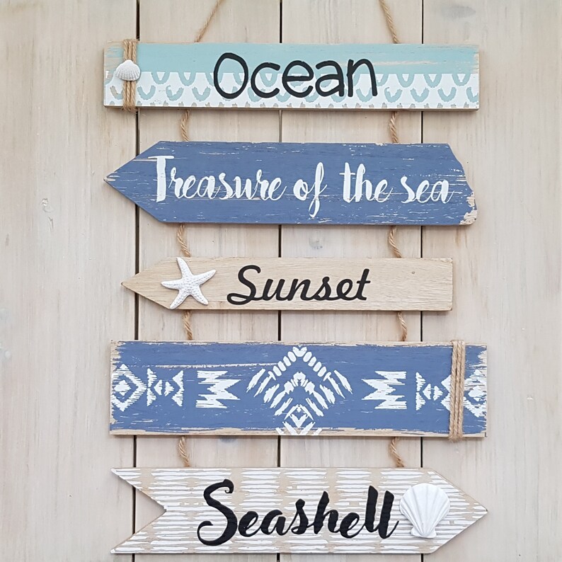 May include: Five wooden signs with a nautical theme hang from twine. The signs are painted in shades of blue, white, and brown. The top sign reads "Ocean", the second sign reads "Treasure of the sea", the third sign reads "Sunset", the fourth sign has a geometric pattern, and the bottom sign reads "Seashell".
