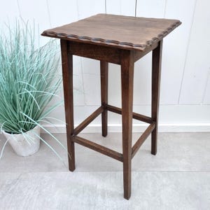 Op de afbeelding: Een hoge, donkerbruine houten bijzettafel met een vierkant blad en geschulpte randen. De tafel heeft vier poten en een onderste draagconstructie. De tafel is ongeveer 76 cm hoog.