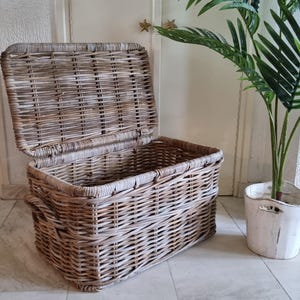 May include: A large, rectangular wicker storage basket with a hinged lid. The basket is made of natural woven rattan and has a light brown colour.