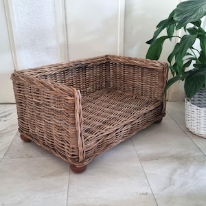 Op de afbeelding: Een rechthoekig huisdierbed gemaakt van gevlochten lichtbruin riet. Het bed heeft hoge zijkanten en een platte bodem, ondersteund door vier kleine, afgeronde houten pootjes. Het vlechtpatroon is consistent door de gehele structuur heen.