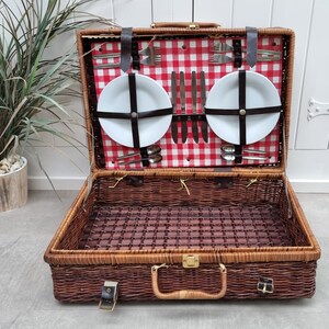 May include: A brown wicker picnic basket with a red and white checkered lining. The basket is open and contains two white plates, silverware, and a set of knives.