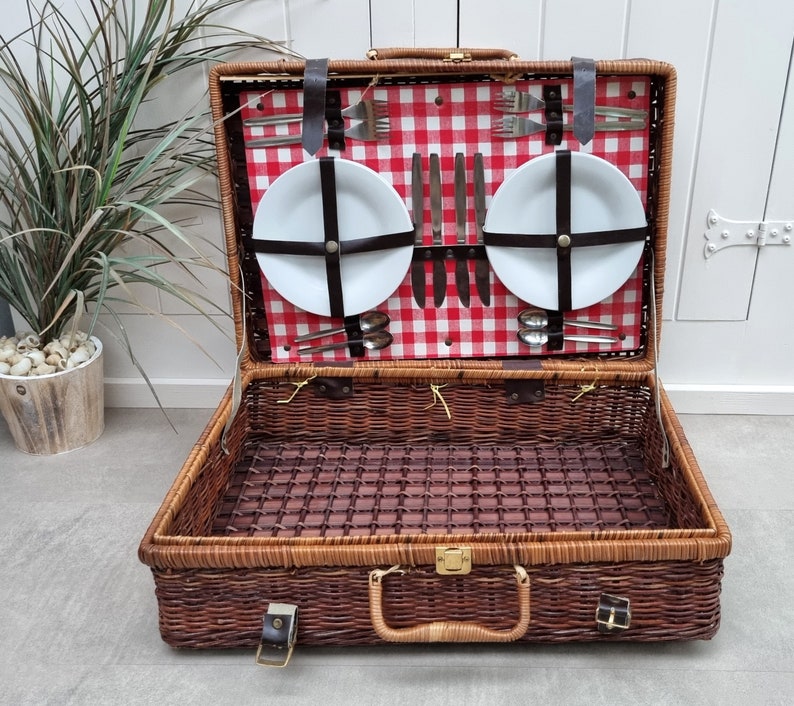 May include: A brown wicker picnic basket with a red and white checkered lining. The basket is open and contains two white plates, silverware, and a set of knives.