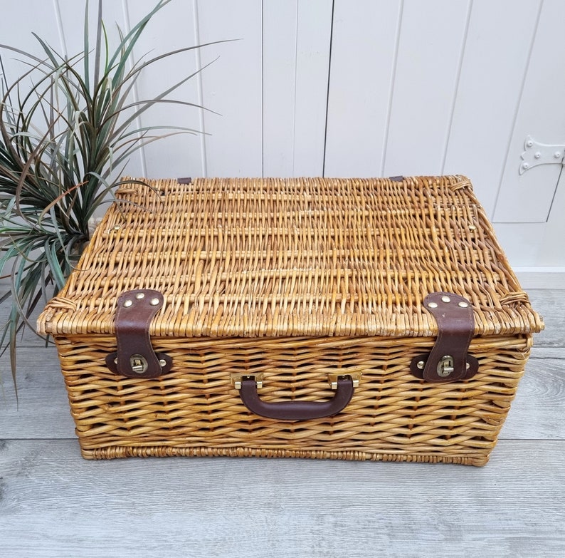 May include: A woven wicker basket with a brown leather handle and brass accents. The basket is closed and has a lid.